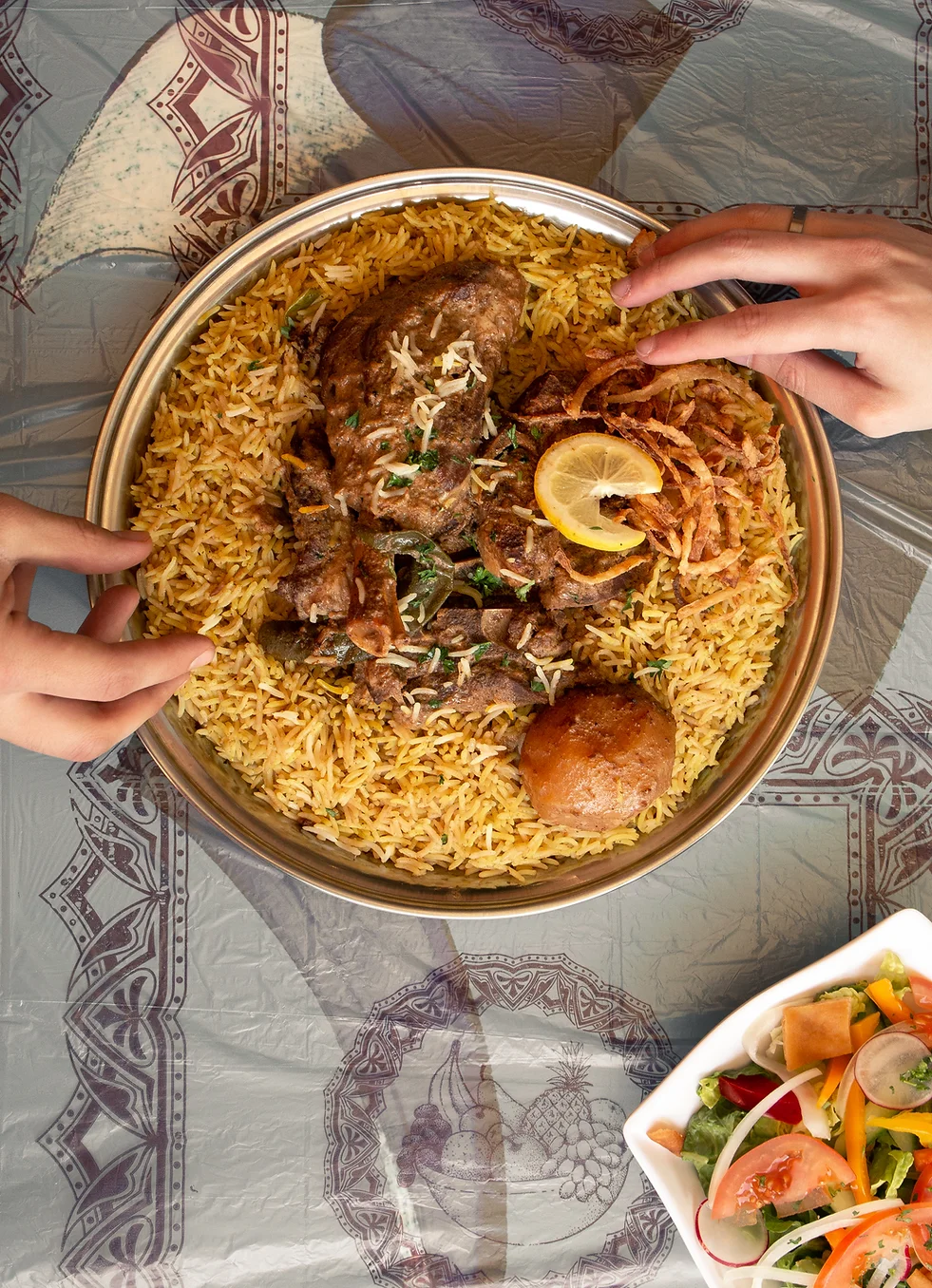 Hands reaching into a golden platter of zurbian rice topped with slow-cooked lamb, crispy onions and lemon.
